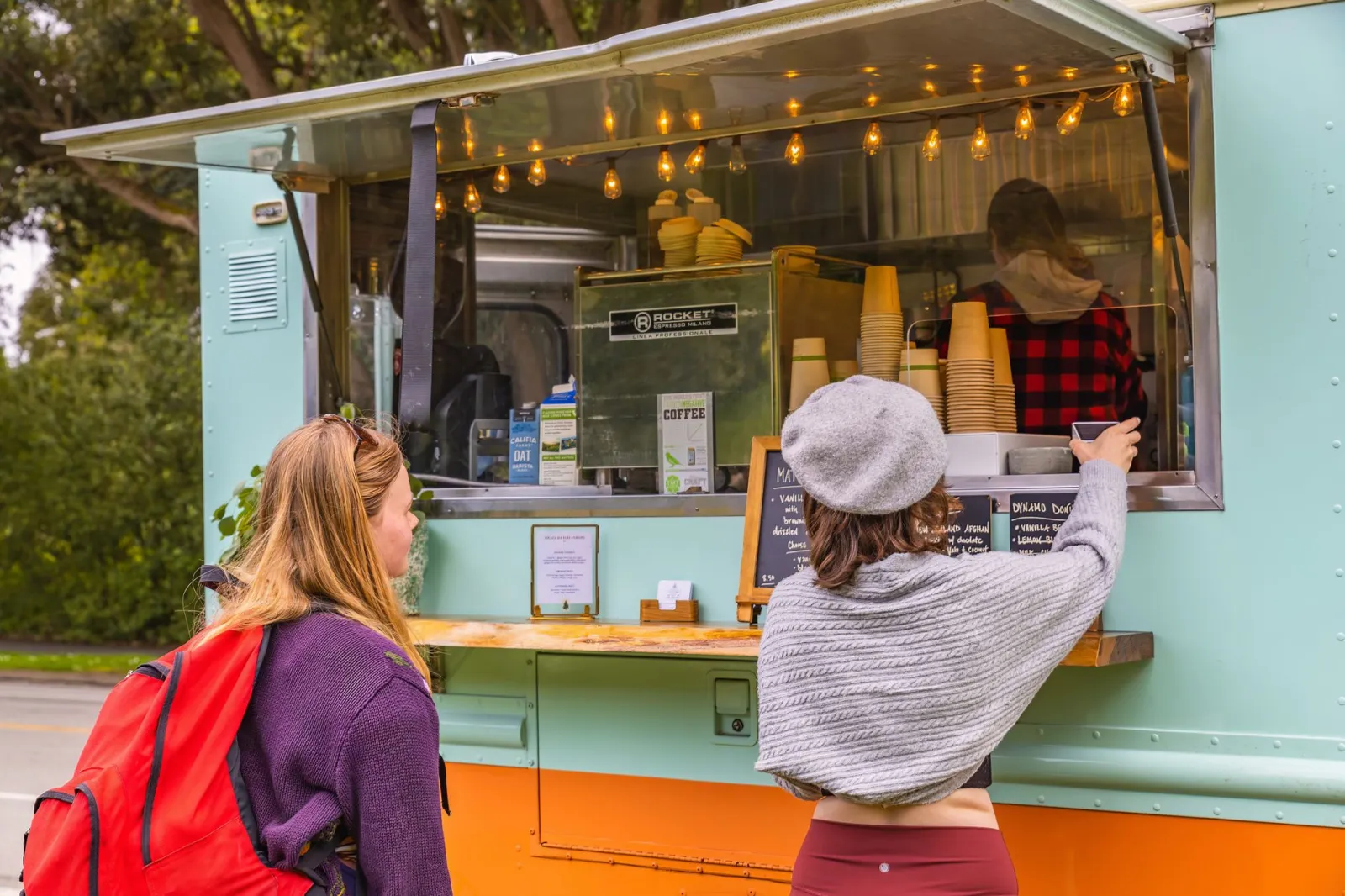 Two women stand at a food truck ordering coffee on a bright day.